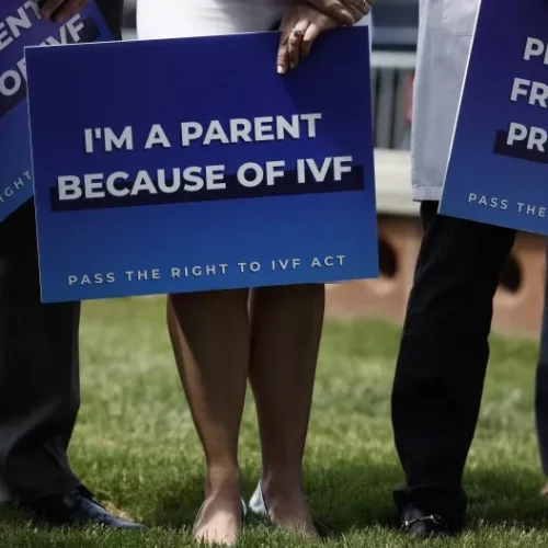 Three people stand on grass holding blue signs supporting IVF; the center sign reads IM A PARENT BECAUSE OF IVF and the others have similar messages. Their faces are not visible.