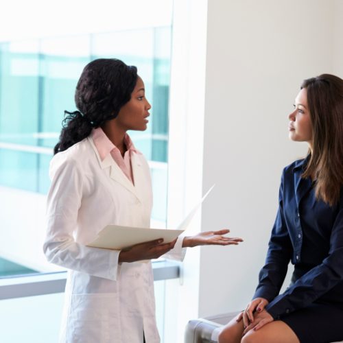 A female doctor in a white coat holding a clipboard talks to a woman sitting on an exam table in a medical office, both appearing engaged in conversation.