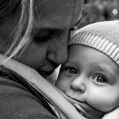 A close-up black and white photo of an adult lovingly holding a baby, who is wearing a knit hat and looking at the camera while nestled in a carrier. The adult’s face is close, showing affection and tenderness.
