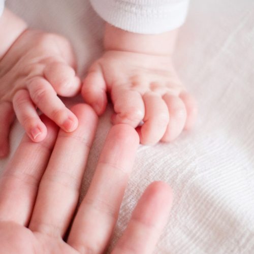 A close-up of an adult hand reaching out, gently holding the tiny hands of a baby, both resting on a soft white blanket.