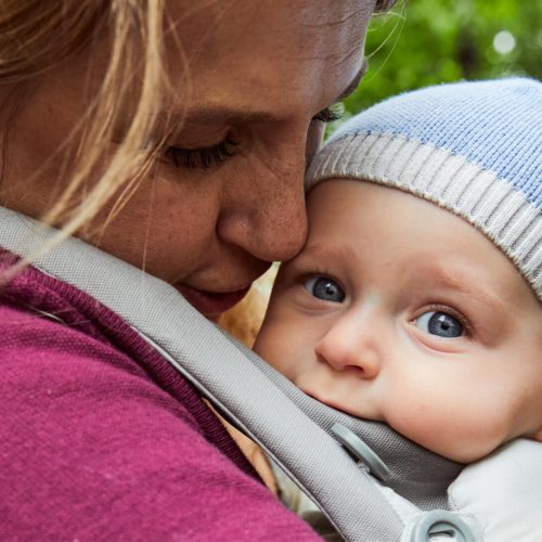 A close-up of an adult gently cuddling a baby in a carrier outdoors. The baby, wearing a blue knit hat, looks ahead with wide blue eyes while the adult’s face is lovingly pressed close to the babys head.