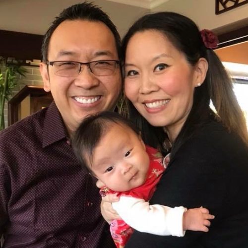 A smiling couple poses together indoors while holding a baby dressed in red. The man wears glasses and a maroon shirt; the woman has long dark hair, styled with a flower, and is wearing a black sweater.