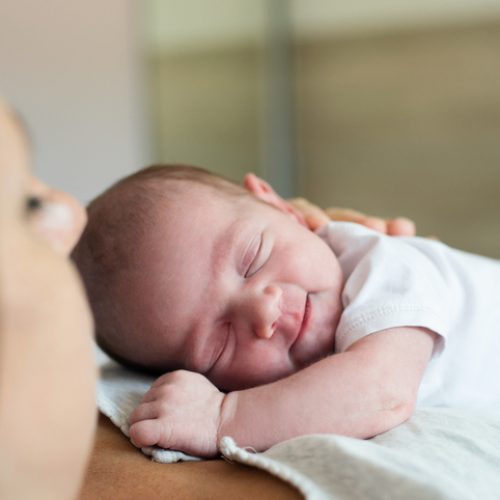 A newborn baby wearing a white onesie sleeps peacefully on an adult’s chest, with the adult’s face partially visible in the foreground. The background is softly blurred.