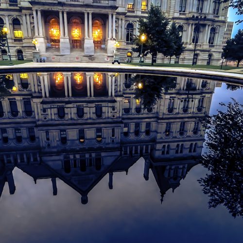 Elegant historic building with glowing lights reflected clearly in a large, still pool of water at dusk, with surrounding trees and part of the sky also visible in the reflection.