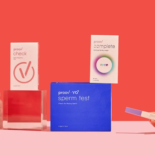 A hand holds a pregnancy test beside various fertility and health products, including Proov check, Proov complete, Proov sperm test, and a bottle of capsules, all arranged on a pink surface with a red background.