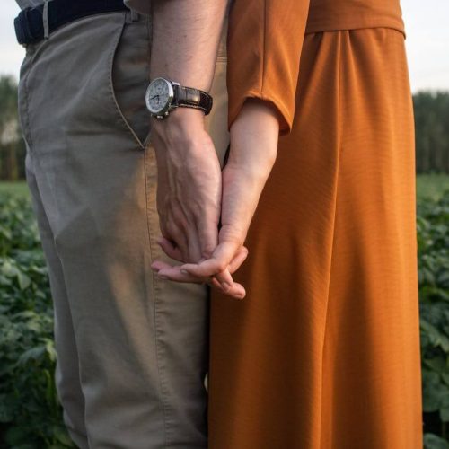 A couple stands close together in a green field, holding hands. One person wears beige pants and a watch, while the other wears an orange dress. Their hands are intertwined, and trees are visible in the background.