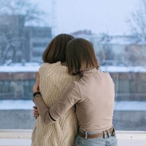 Two people with short brown hair sit close together by a large window, hugging and looking outside at a snowy, blurred cityscape. They appear to be offering comfort to each other.