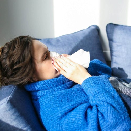 A person wearing a blue sweater sits on a couch and holds a tissue to their nose, appearing to sneeze or blow their nose. Blue cushions are in the background, and sunlight comes through a nearby window.