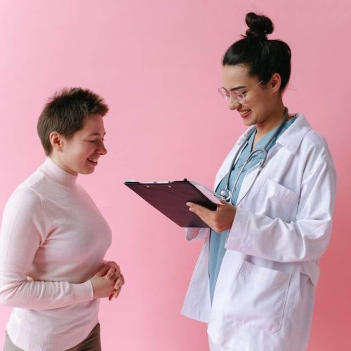 A doctor in a white coat and stethoscope smiles while holding a clipboard, speaking with a patient who is also smiling, against a pink background.