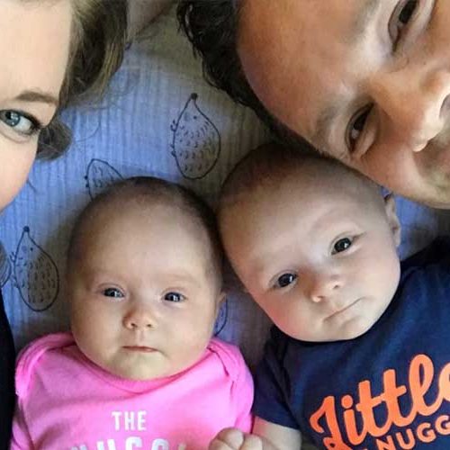A smiling woman and man lie next to two babies. The baby on the left wears a pink onesie, and the baby on the right wears a blue onesie that says Little Nugget. They are all lying close together, looking at the camera.