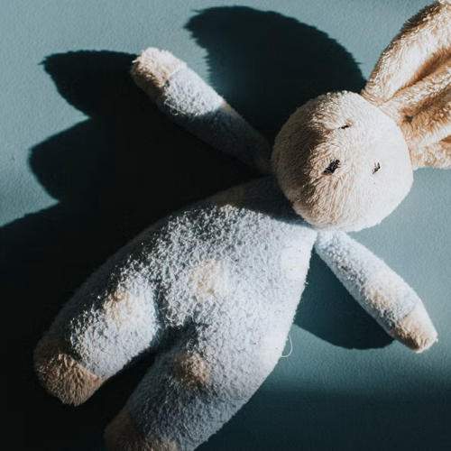 A plush bunny toy with long ears lies on a blue surface, casting a shadow to the left. The bunny appears soft and slightly worn, with light cream and blue coloring.