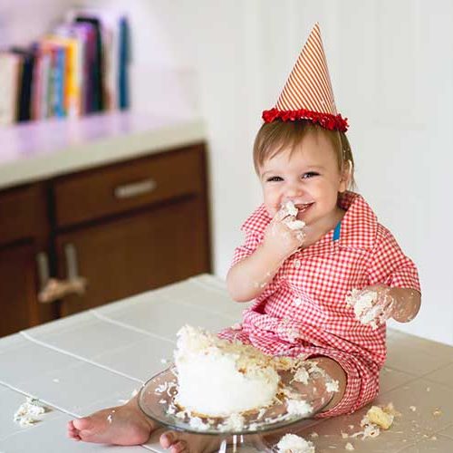 A smiling toddler in a red checkered outfit and party hat sits on a kitchen counter, happily eating and playing with a smashed birthday cake, with cake smeared on their face and hands.