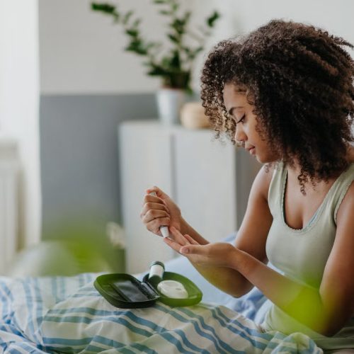 A woman with curly hair sits on a bed, using a blood glucose meter to check her blood sugar. She holds a test strip to her finger, with diabetes supplies on a tray in front of her.