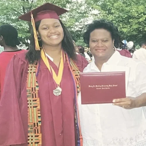 A student in a maroon graduation cap and gown stands smiling next to a woman in white holding a diploma. They are outdoors with other people and trees in the background.