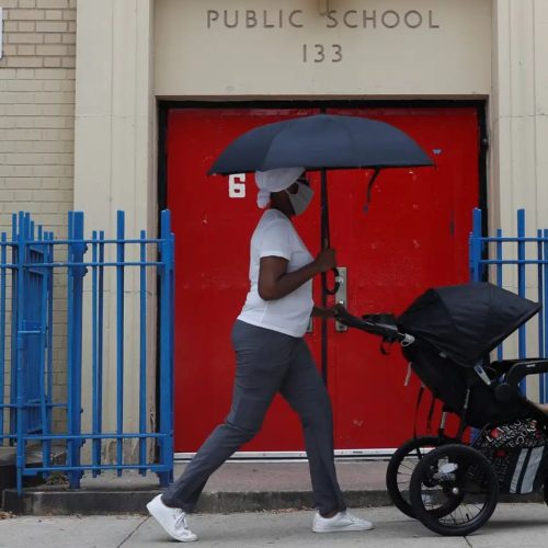 A person wearing a mask walks past a red door at Public School 133, pushing a stroller and holding a black umbrella. Blue metal fencing lines the sidewalk.