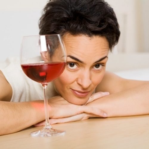 A person with short dark hair rests their chin on their hands on a table, looking at the camera. A glass of red wine sits on the table in front of them.