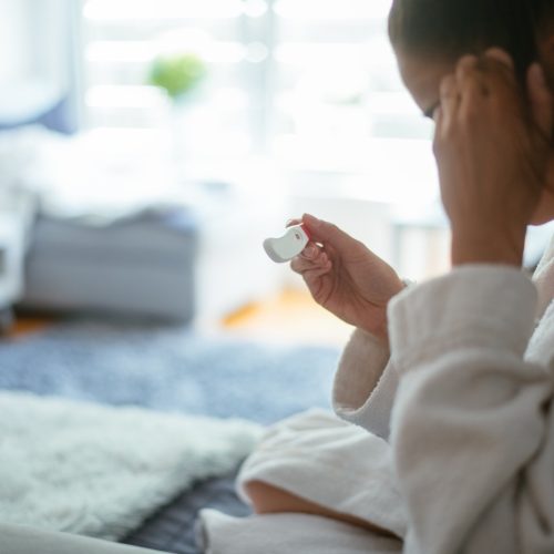A person in a white robe sits indoors, holding a white electronic device and touching their forehead, with a blurred living room in the background.