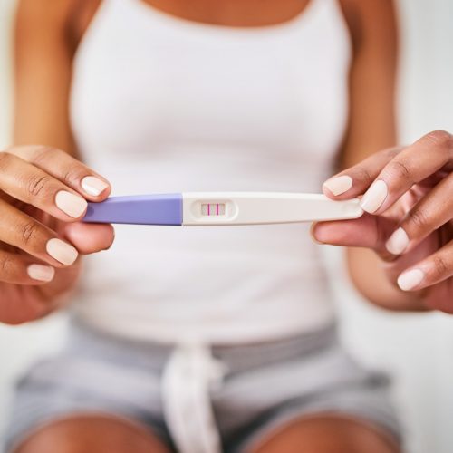 A person in casual clothes holds a pregnancy test showing two pink lines, indicating a positive result. The image focuses on the test and their hands.