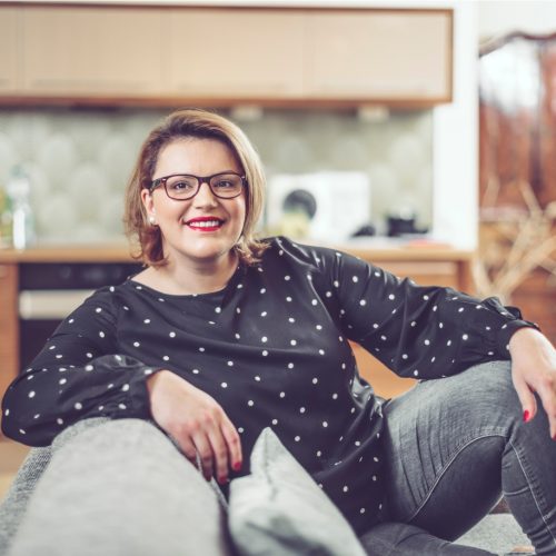 A woman with glasses and shoulder-length hair, wearing a black polka dot top and gray jeans, sits comfortably on a couch in a modern kitchen, smiling at the camera.