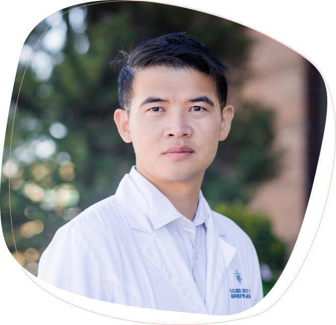 A man with short black hair, wearing a white lab coat and collared shirt, stands outdoors with greenery and a building in the blurred background.